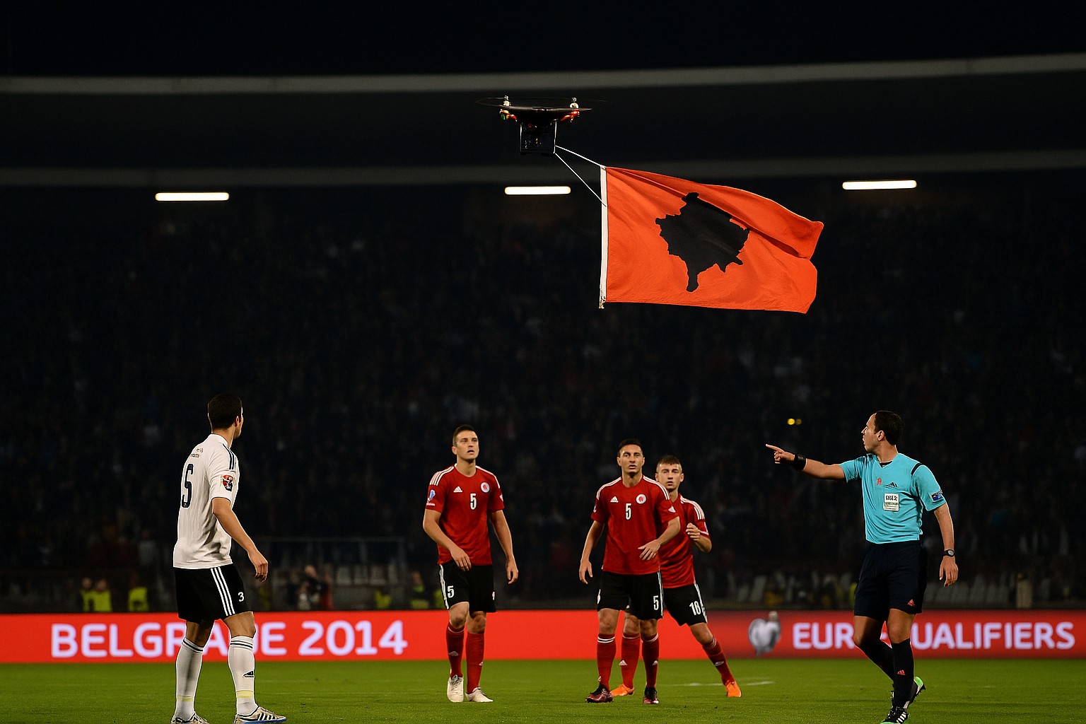 Ein Fußballspiel unter Flutlicht wird jäh unterbrochen, als eine Drohne mit einer Flagge der „Großalbanien“-Silhouette über das Spielfeld fliegt. Spieler beider Teams und der Schiedsrichter blicken fassungslos in die Luft, während das Stadion im Hintergrund brodelt.
